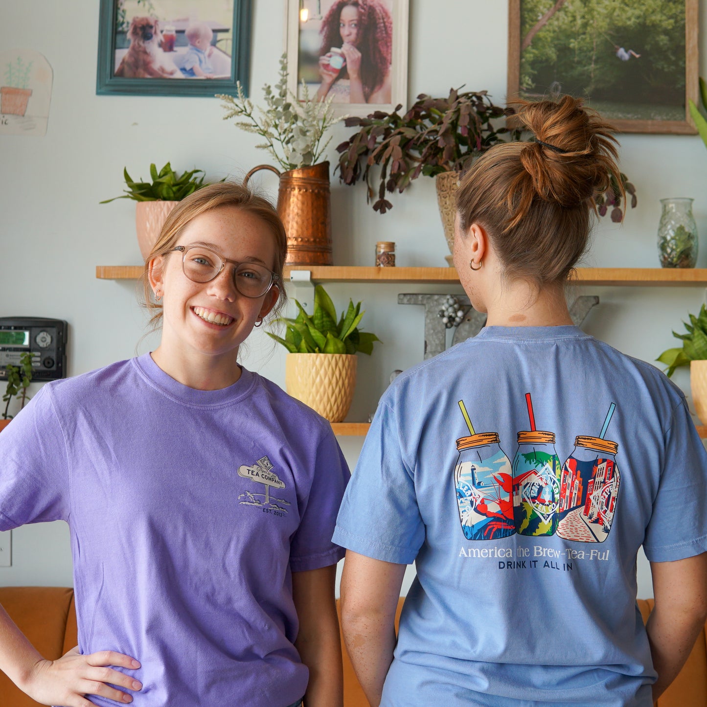 Two women are in a room with potted plants; one smiles at the camera in lavender, while the other faces away, wearing a blue Piper and Leaf Tea Co. Nor'Easter Adventure T-shirt featuring mason jars and straws.