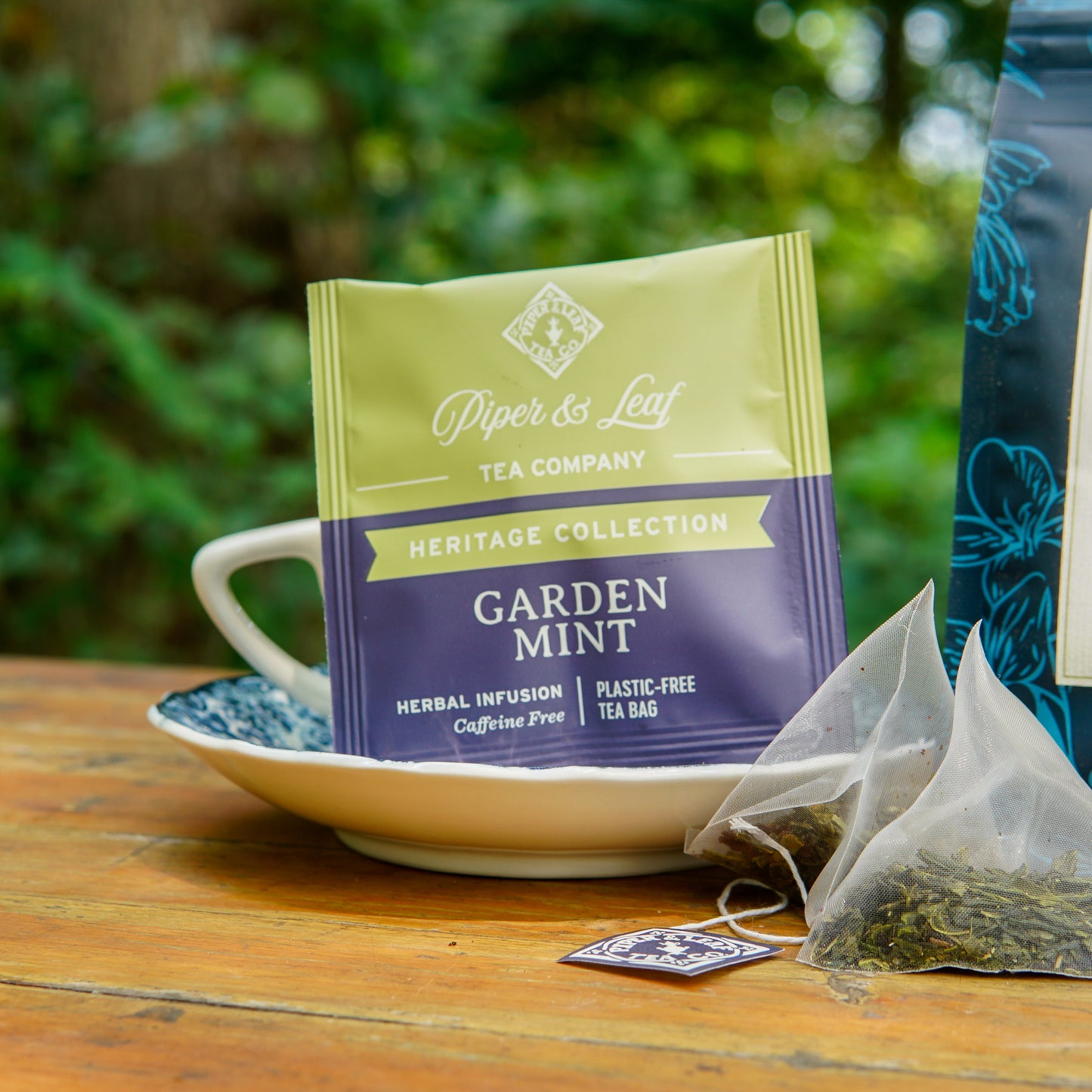 A Piper and Leaf Tea Co. Garden Mint 50ct Individually Wrapped Tea Bag rests on a teacup and saucer, with loose herbs scattered nearby atop a wooden outdoor table. This refreshing herbal blend is entirely caffeine free—ideal for peaceful garden relaxation, much like a classic peppermint chamomile tea.
