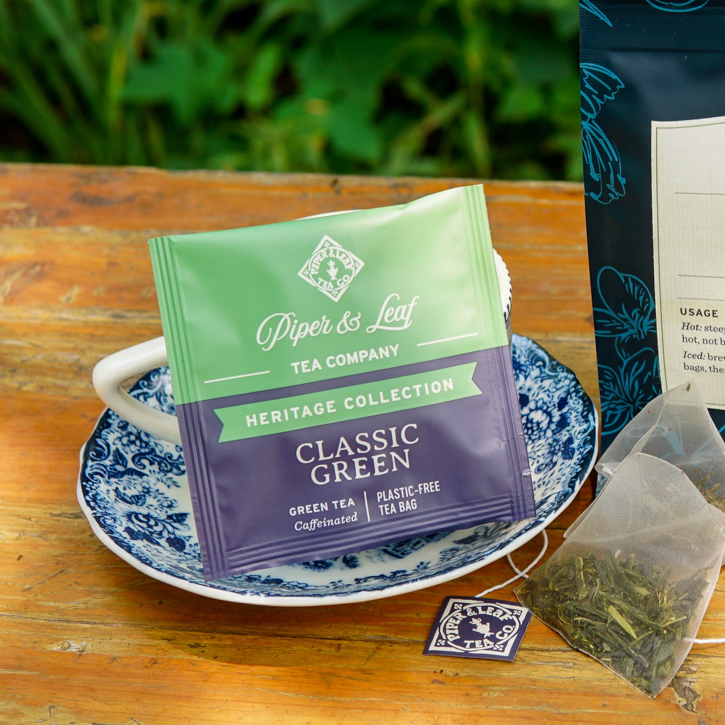 A Piper and Leaf Tea Co. Classic Green 50ct Individually Wrapped Tea Bag, made from plastic-free materials, rests on a blue-and-white saucer alongside a cup, a used tea bag, and the tea package on a wooden surface with greenery in the background.