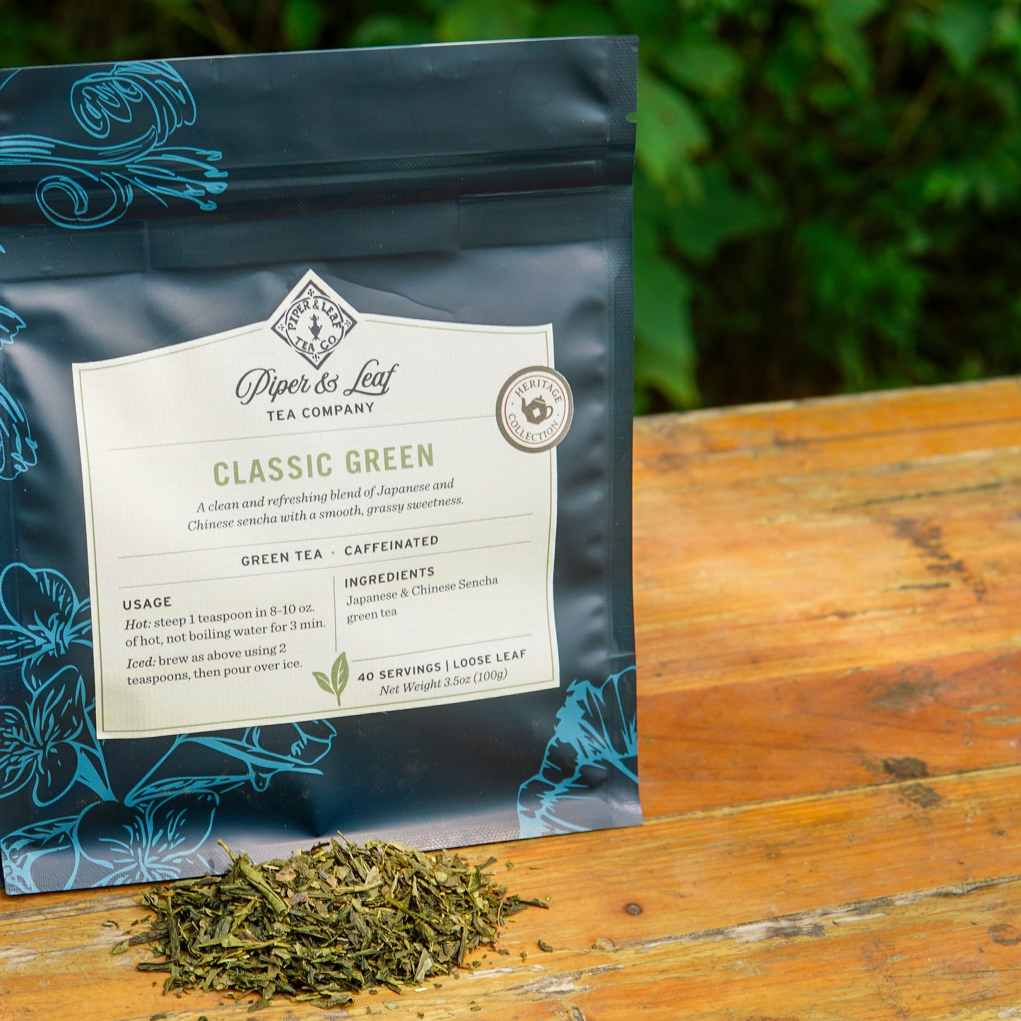 A package of Piper and Leaf Tea Co. Classic Green 40 Serving Loose Leaf tea rests on a wooden surface outdoors, with a small pile of sencha green tea leaves displayed in front.