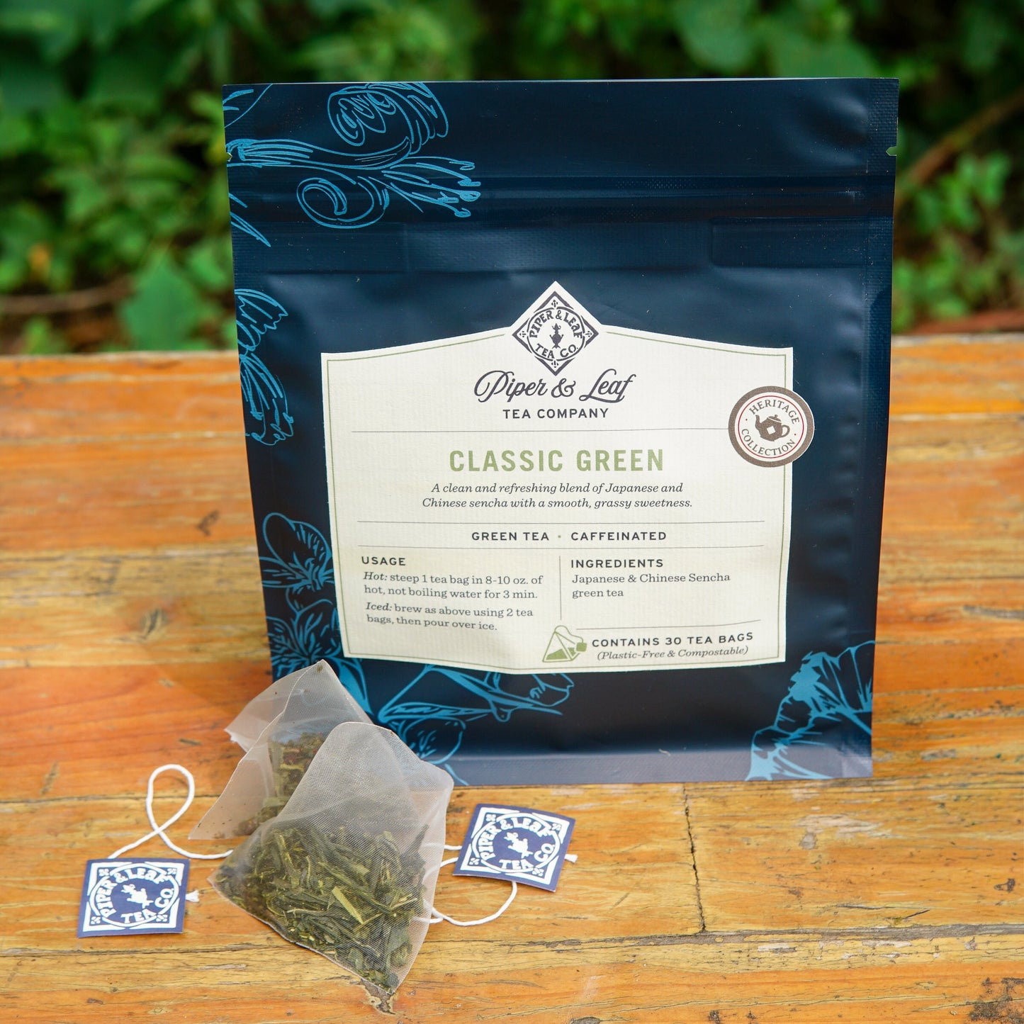 A box of Piper and Leaf Tea Co. Classic Green 30ct Tea Bags is showcased on a wooden surface with two plastic-free tea bags placed in front, while blurred green foliage appears in the background.