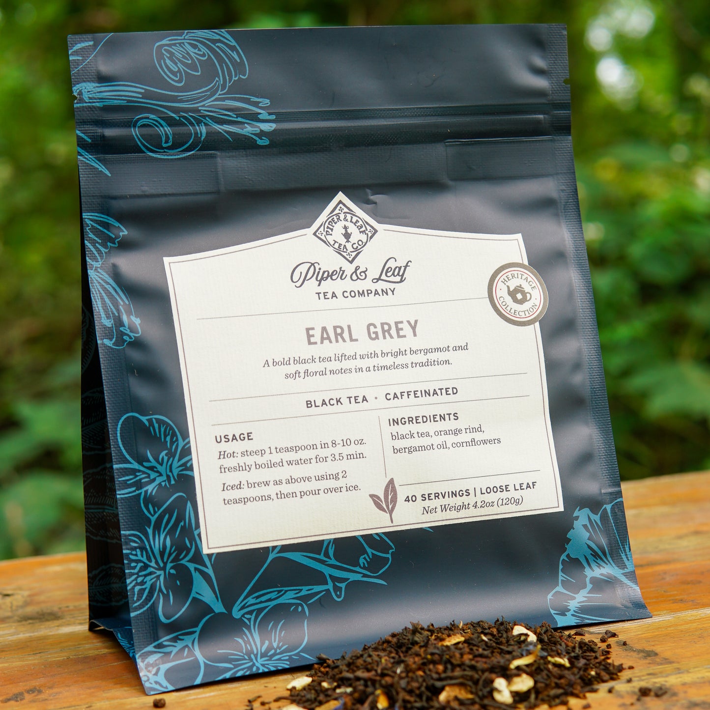 A bag of Piper and Leaf Tea Co.’s Earl Grey 40 Serving Loose Leaf Tea is placed outdoors on a wooden surface, with black tea leaves scattered in front of it.