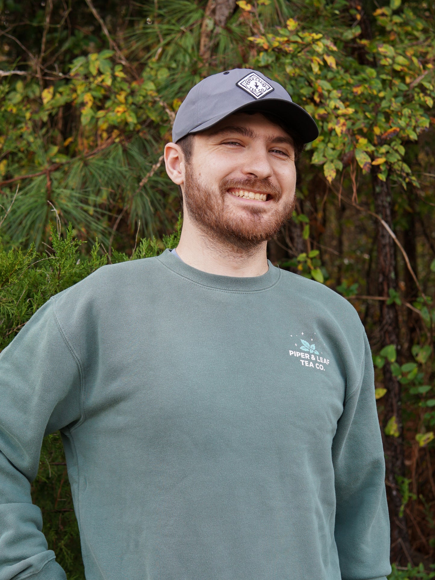 A bearded man in a âPiper and Leaf Tea Co.â It Came Upon a Minty Midnight Sweatshirt and gray cap smiles outdoors by leafy trees, perhaps savoring the fresh vibe of this cozy, caffeine-free inspired look.