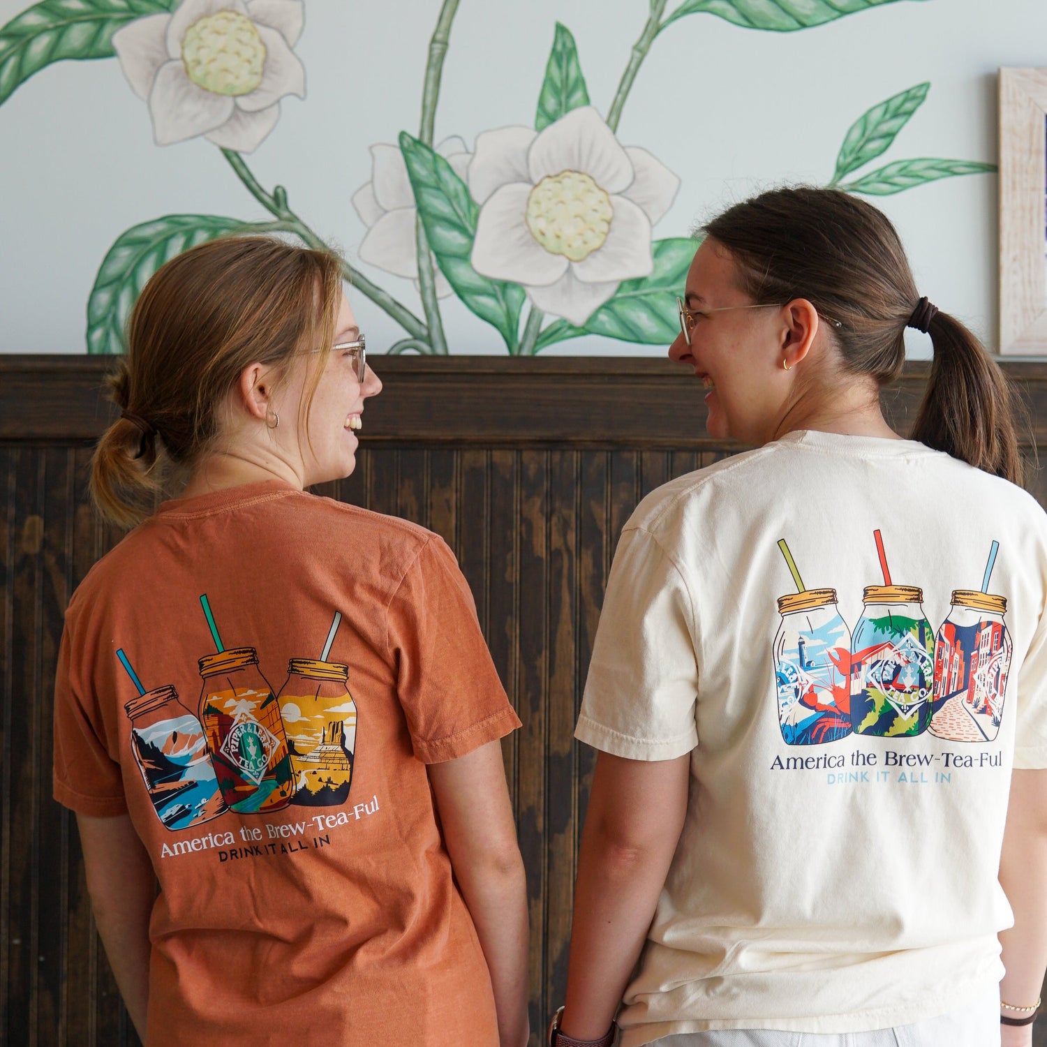 Two people stand side by side, smiling at each other. Both wear T-shirts with colorful mason jar graphics and the text "America the Brew-Tea-Ful." A floral mural is on the wall behind them.
