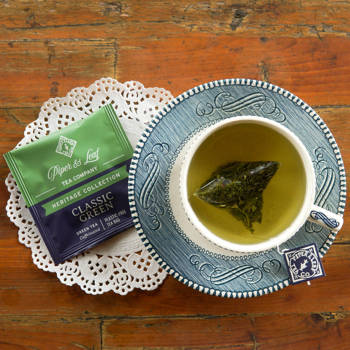 A cup of green tea with a tea bag, a packet labeled “Classic Green” by Piper & Leaf Tea Co., and a paper doily on a wooden table.