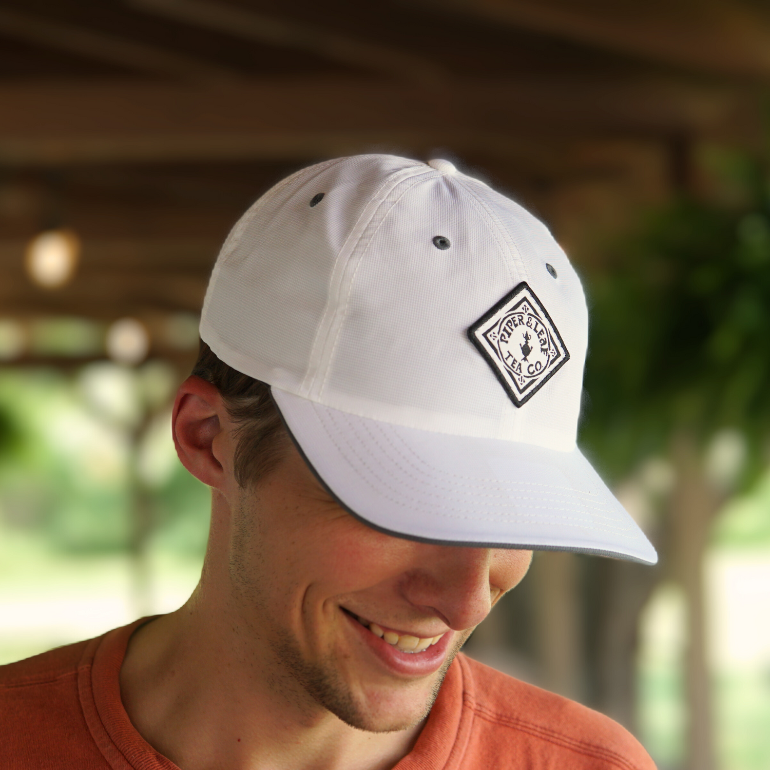 A man wearing a white baseball cap with a black and white patch on the front looks down and smiles. He is dressed in an orange shirt, and the background is blurred outdoors.