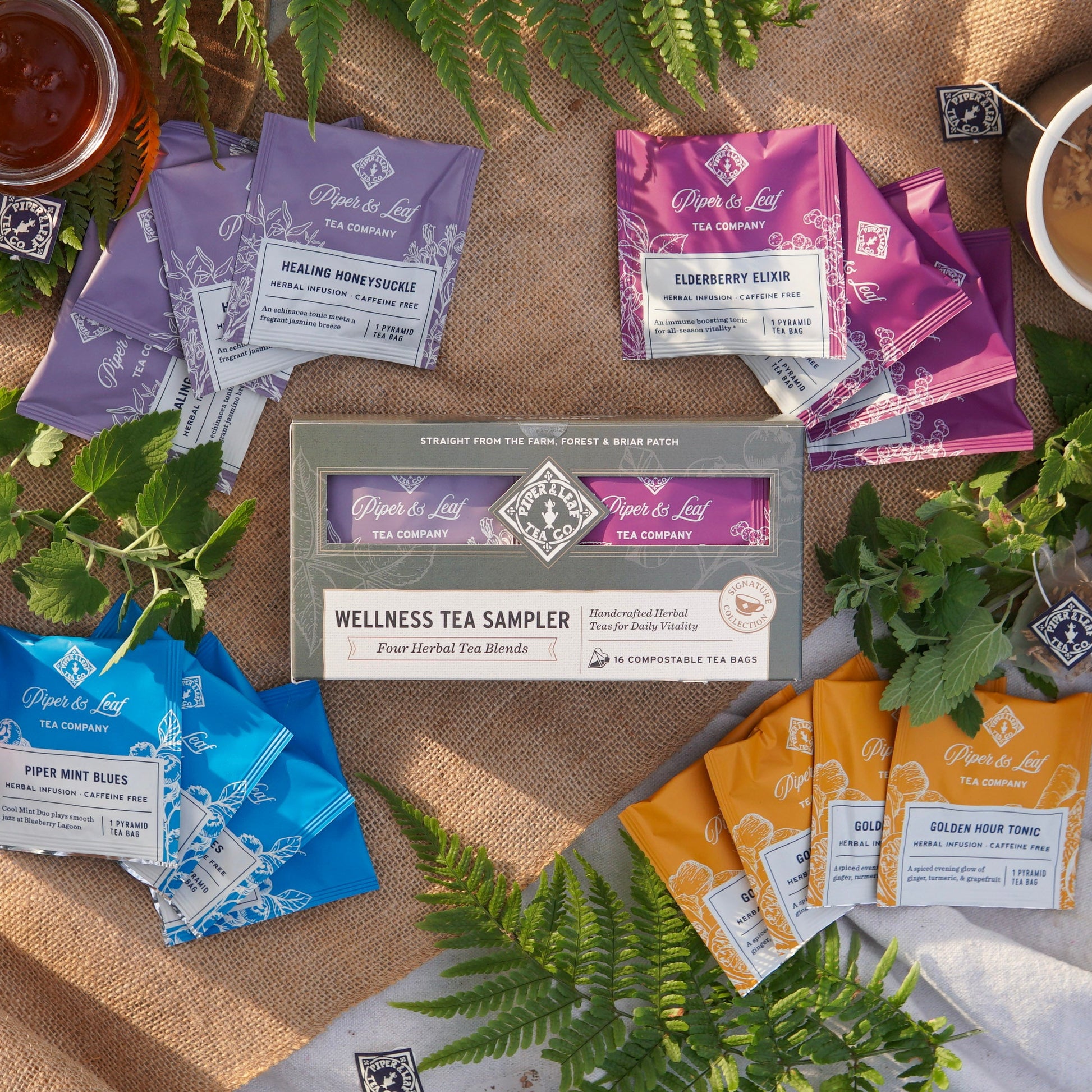 Piper & Leaf Tea Co. Wellness Tea Sampler – Box of 16 tea bags is displayed on burlap, surrounded by loose herbal infusion tea, cups, and tea labels.