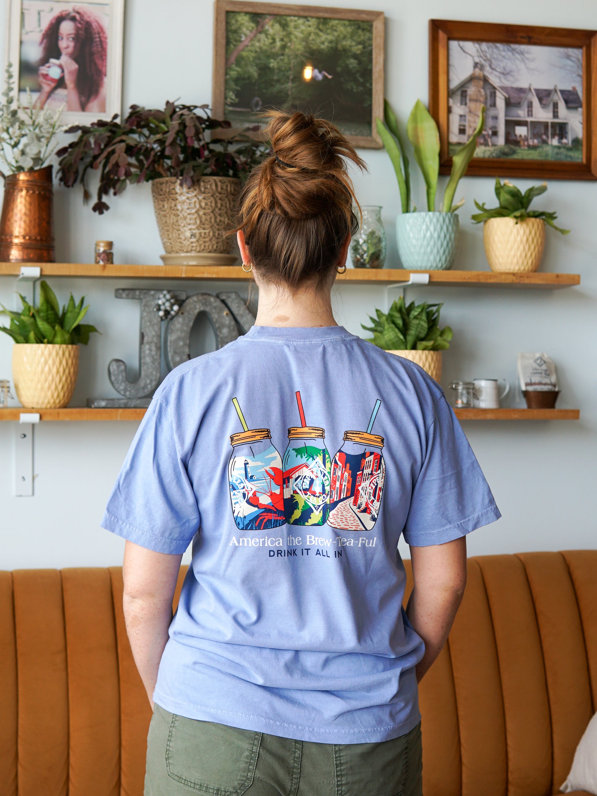 Indoors, a person faces a wall of plants, wearing the Piper and Leaf Tea Co. Nor'Easter Adventure T-shirt featuring three mason jars and the phrase “America the Brewtiful, Drink It All In” on the back.