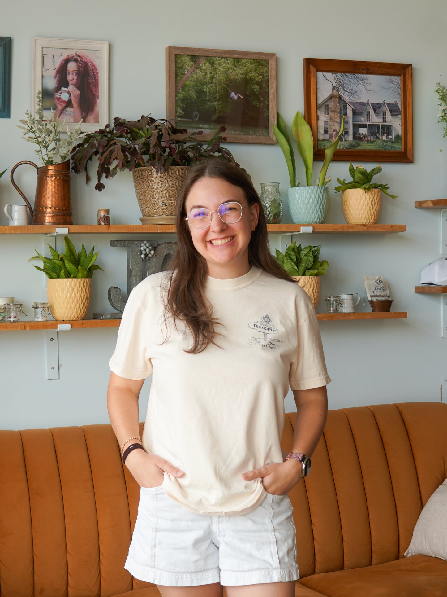 A smiling woman in glasses wears a Nor'Easter Adventure T-shirt by Piper and Leaf Tea Co., standing in a living room with plants, framed photos, and a brown couch.