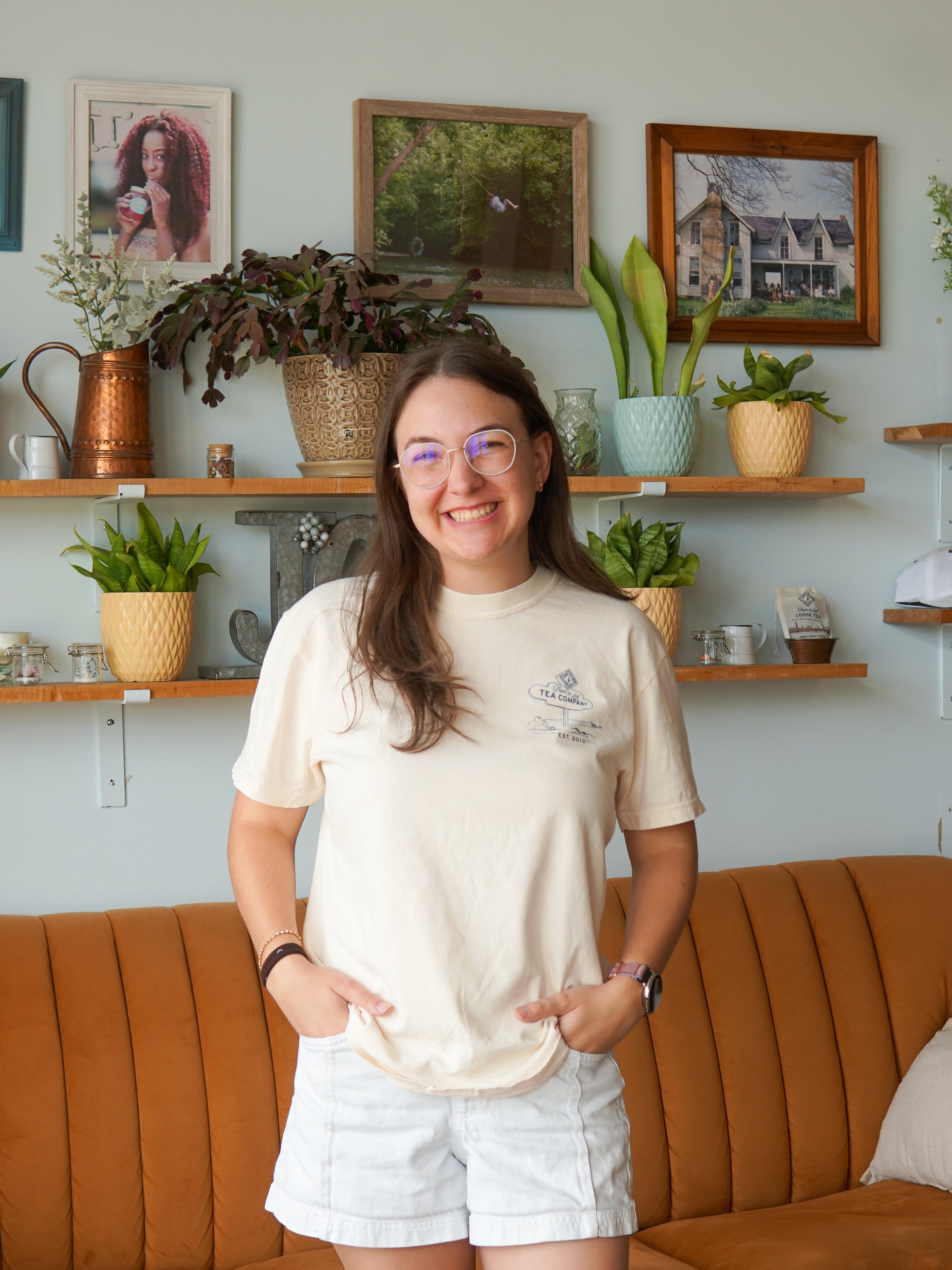 A smiling woman in glasses wears a Nor'Easter Adventure T-shirt by Piper and Leaf Tea Co., standing in a living room with plants, framed photos, and a brown couch.