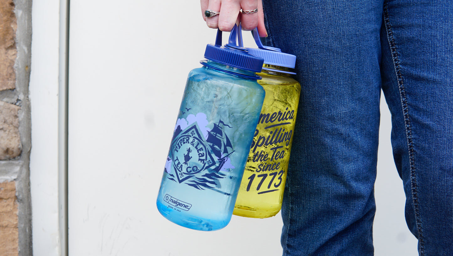 A person in jeans holds a blue bottle filled with ice and a Piper and Leaf Tea Co. "Spilling the Tea" Nalgene yellow water bottle with printed text.
