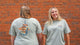 Two women pose against a brick wall. One smiles at the camera while the other shows off the back of her Piper and Leaf Tea Co. Spilling the Tea Shirt, which features a mason jar and the text 