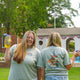 Two women stand outdoors with drinks, both in light green shirts. One wears the 