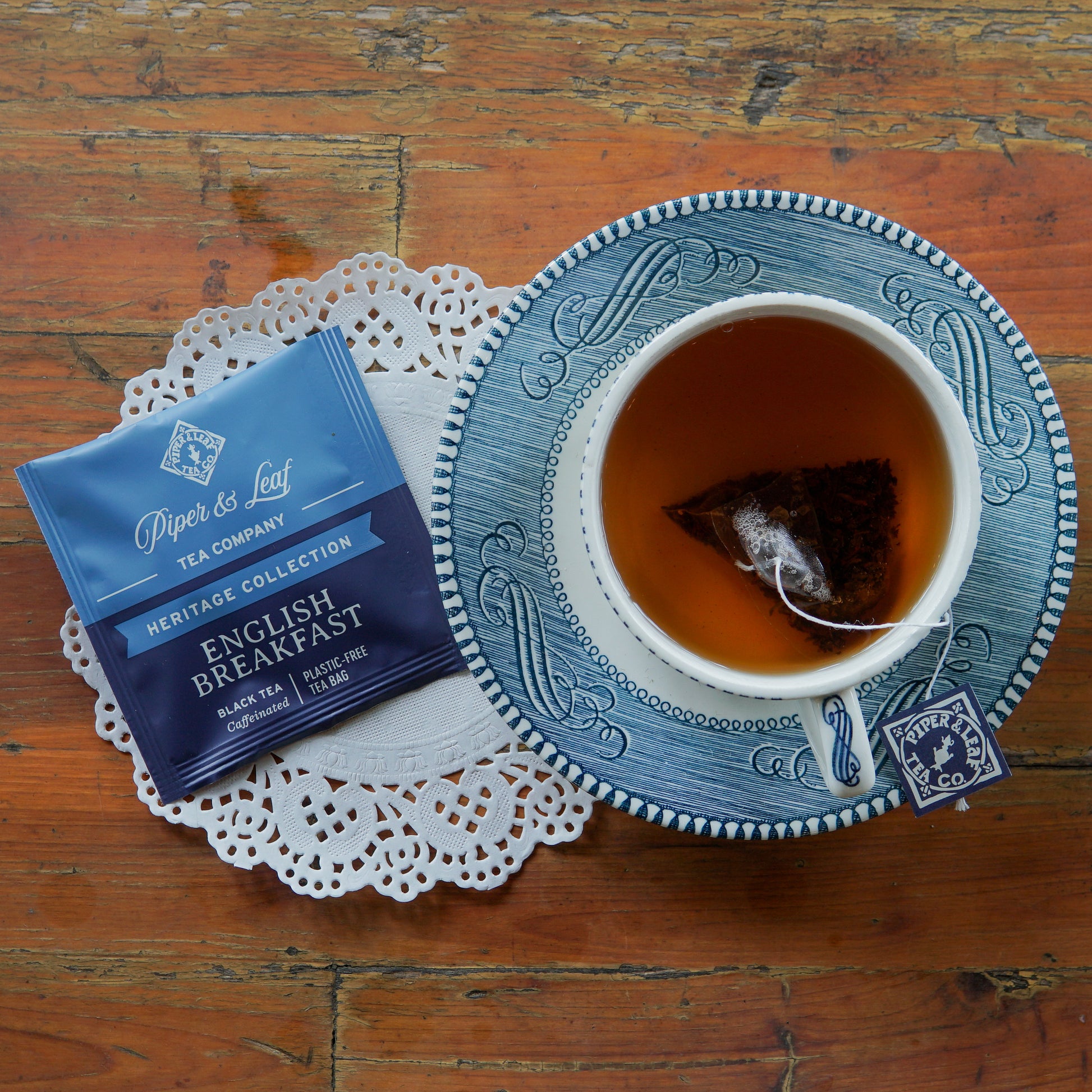 A blue and white teacup filled with tea rests on a saucer and lace doily beside a packet of Piper and Leaf Tea Co. English Breakfast 50ct Individually Wrapped Tea Bags on a wooden table.