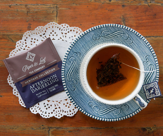 A cup of tea with a tea bag inside sits on a blue saucer next to a packet labeled “Afternoon Darjeeling 50ct Individually Wrapped Tea Bags” from Piper and Leaf Tea Co., displayed on a white doily to showcase the rich flavor of Darjeeling black tea.