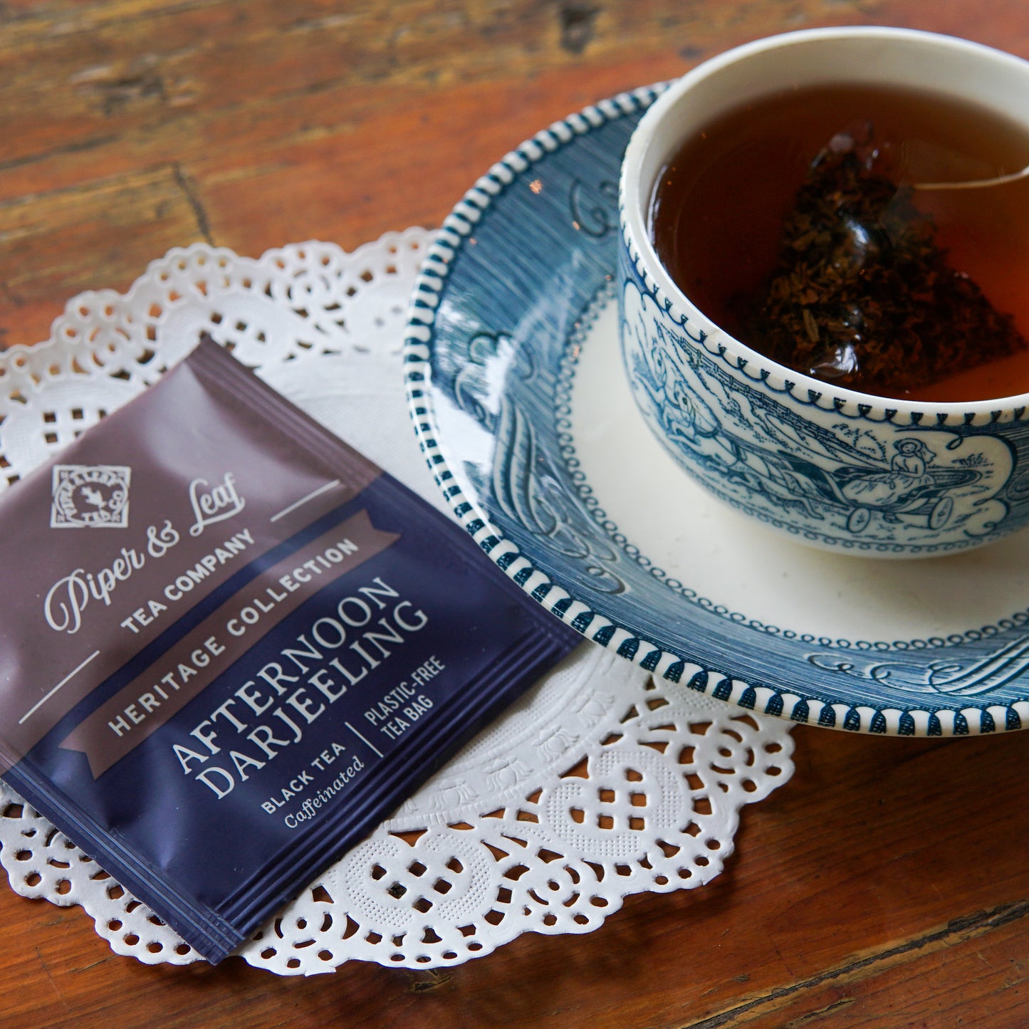 A cup of tea with a tea bag rests on a blue saucer beside a Piper and Leaf Tea Co. Afternoon Darjeeling 50ct Individually Wrapped Tea Bags packet from the Heritage Collection, placed on a lace doily over a wooden table.