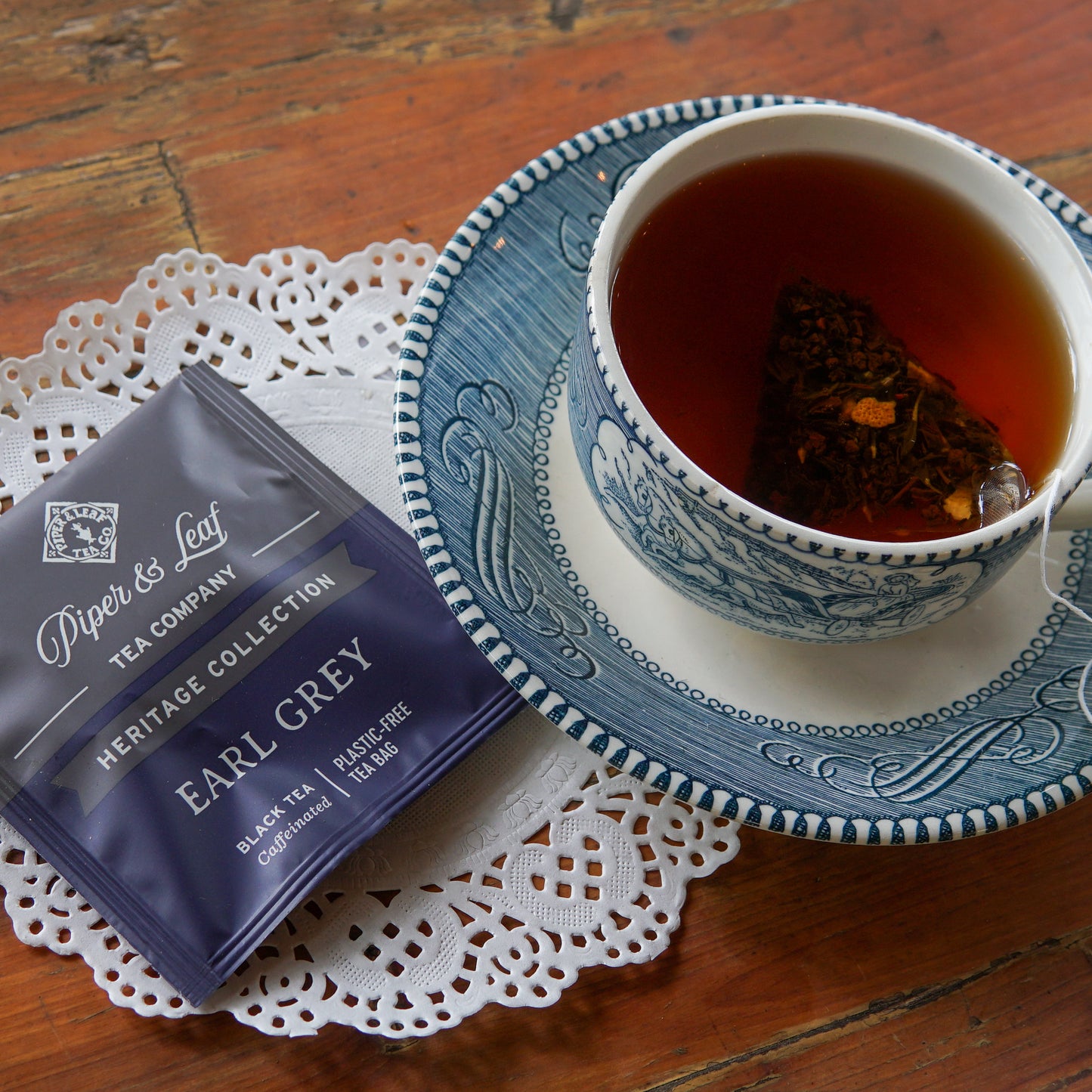 A blue and white teacup filled with tea rests on a saucer adorned with a paper doily, beside a Piper and Leaf Tea Co. Earl Grey 50ct Individually Wrapped Tea Bag, all set atop a wooden table.