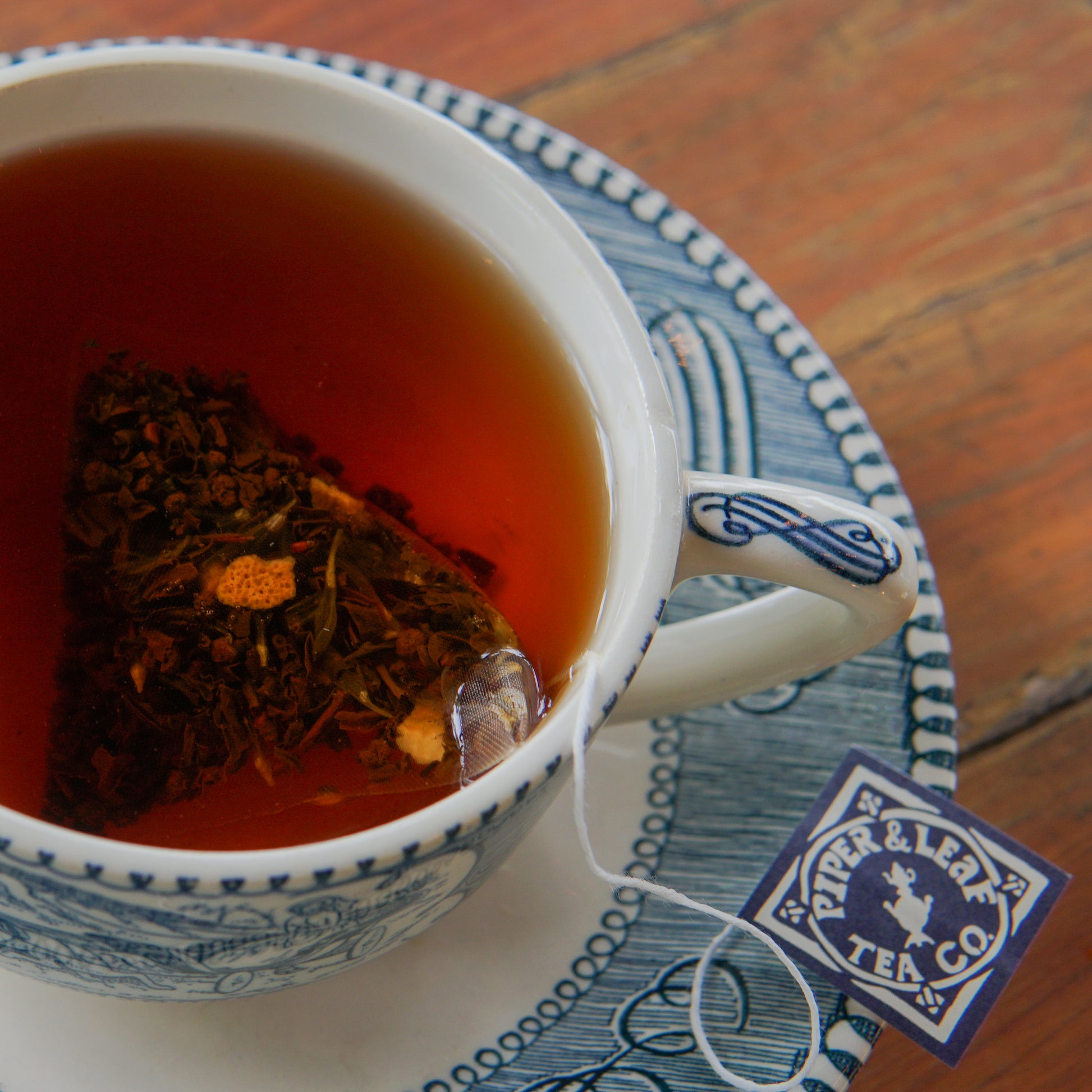 A cup of Earl Grey tea, brewed with a triangular bag from the Earl Grey 30ct Tea Bags by Piper and Leaf Tea Co., sits on a blue and white patterned saucer. The bag’s label, bearing the Piper and Leaf Tea Co. name, hangs over the edge of the cup.