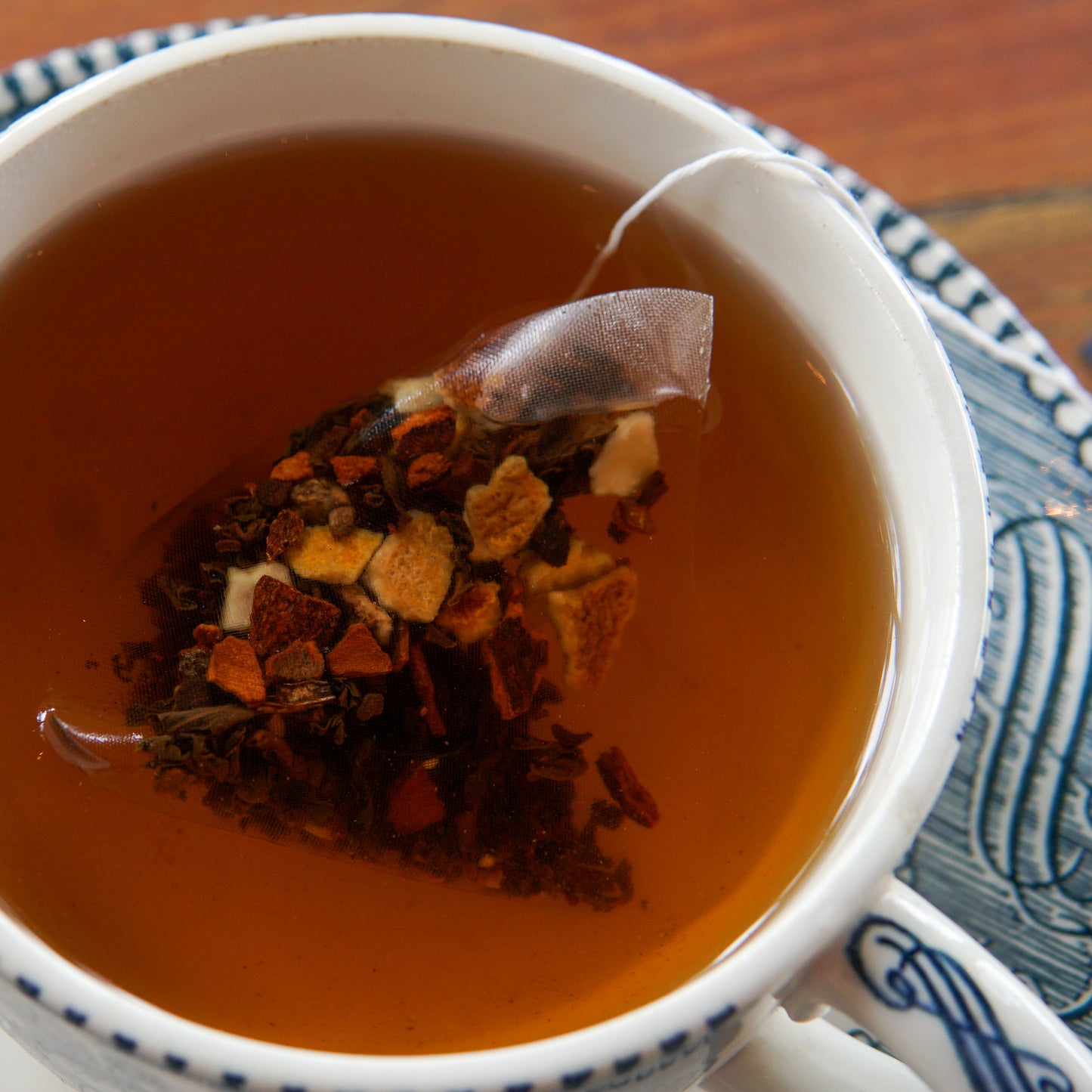 A close-up of a cup of Piper and Leaf Tea Co.’s Cinnamon Orange 30ct Tea Bags from the Heritage Collection, with a pyramid-shaped tea bag steeping amid visible dried fruit and tea leaves, served on a patterned saucer.