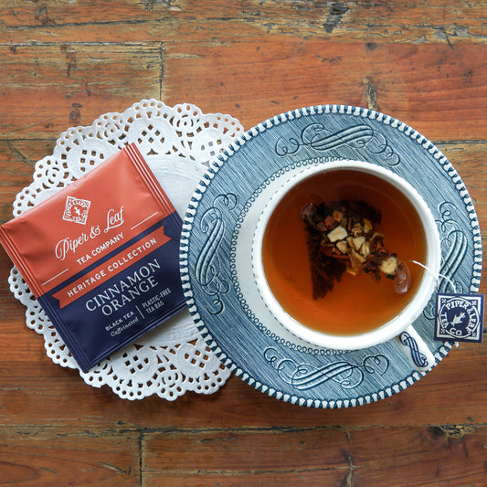 A cup of tea with floating spices rests on a blue saucer beside a box of Piper and Leaf Tea Co. Cinnamon Orange 50ct Individually Wrapped Tea Bags, set on a doily atop a wooden table.