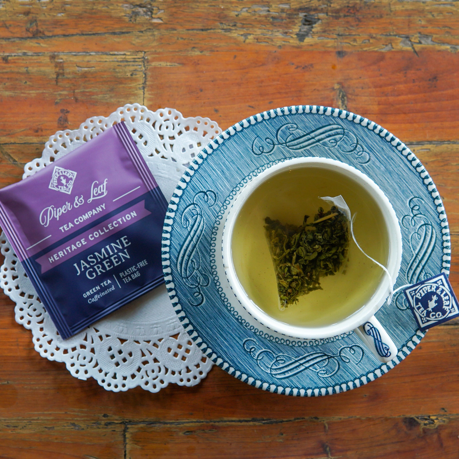 A cup of Piper and Leaf Tea Co. Jasmine Green tea, brewed from a plastic-free, individually wrapped tea bag, rests on a blue-and-white saucer beside a purple packet from the Heritage Collection. The ensemble sits atop a white doily on a wooden table.