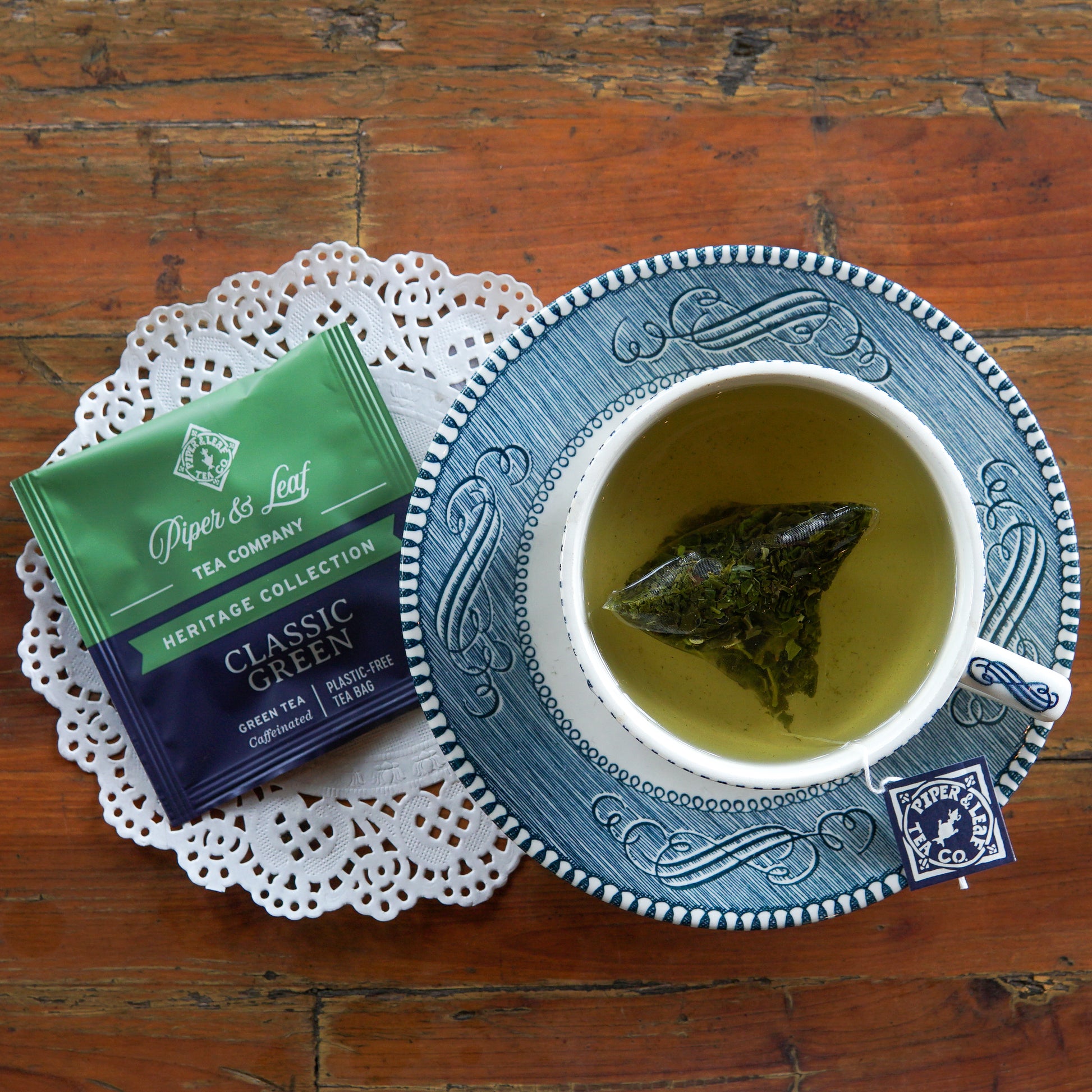 A cup of green tea with a plastic-free tea bag steeping, next to a packet labeled “Piper and Leaf Tea Co. Classic Green 50ct Individually Wrapped Tea Bags,” sits on a lace doily atop a wooden table.