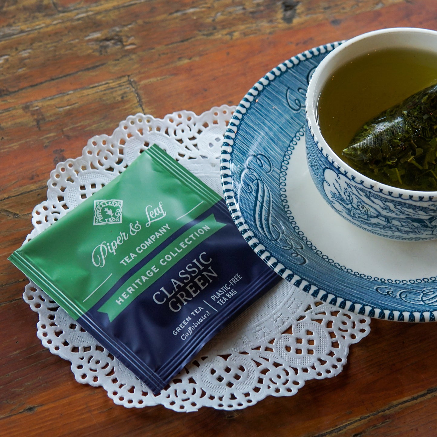 A packet of Piper and Leaf Tea Co.’s Classic Green 50ct Individually Wrapped Tea Bags rests on a lace doily beside a blue and white teacup filled with sencha green tea, showcasing its plastic-free tea bags.