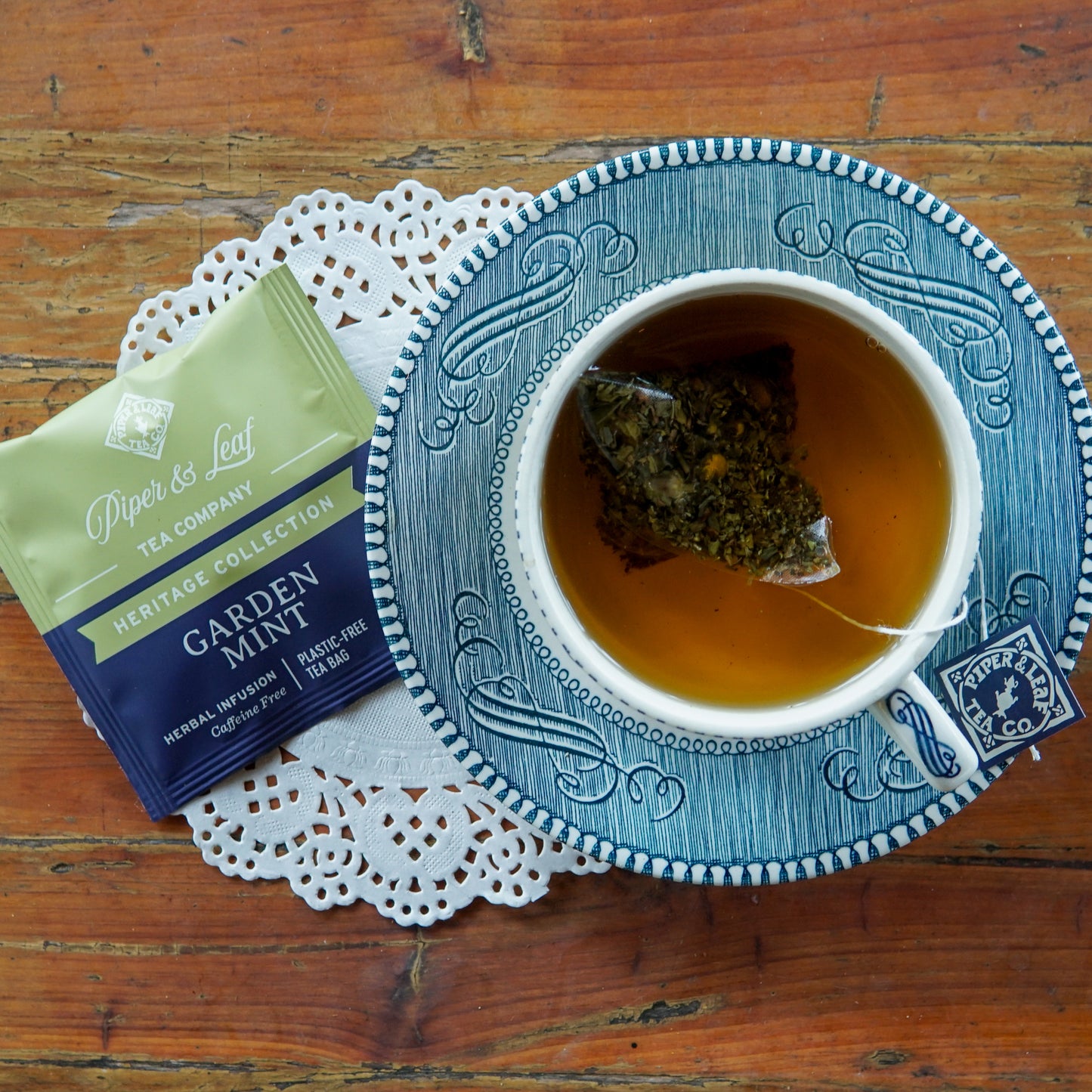 A cup of tea with a visible tea bag rests on a blue-patterned saucer beside a Piper and Leaf Tea Co. “Garden Mint 50ct Individually Wrapped Tea Bags” packet, all set on a lace doily atop a wooden surface, inviting you to savor this naturally caffeine-free and refreshing herbal infusion.