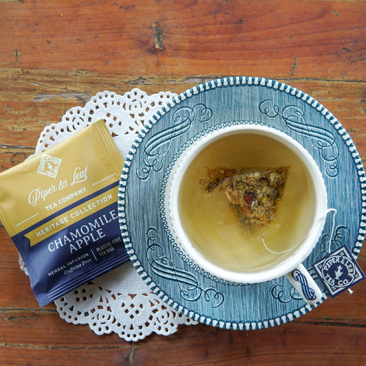 A cup of Piper and Leaf Tea Co. Chamomile Apple tea, a soothing caffeine-free herbal infusion with an individually wrapped tea bag inside, sits on a blue saucer next to a packet of Chamomile Apple 50ct Individually Wrapped Tea Bags on a lace doily atop a wooden table.