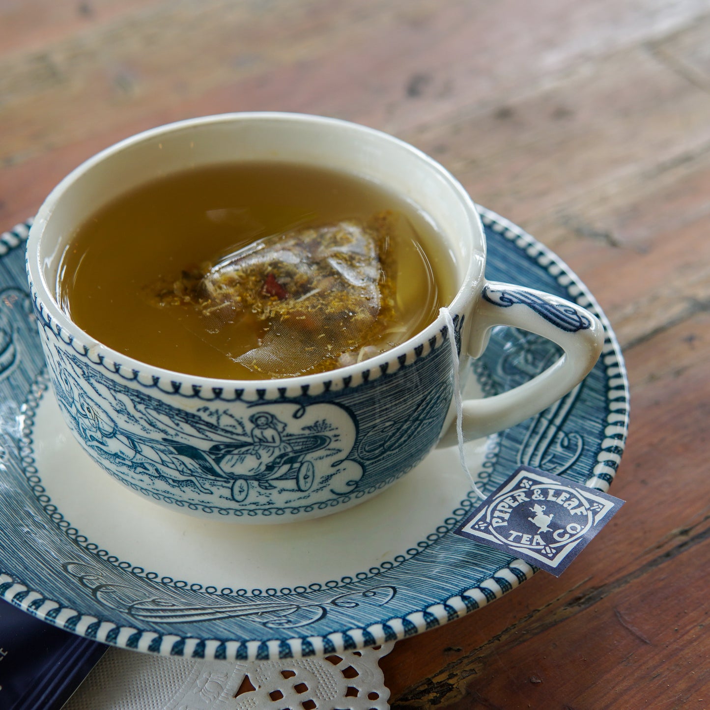 A patterned teacup filled with hot tea and a tea bag sits on a matching saucer on a wooden table. The tea bag label reads “Chamomile Apple 30ct Tea Bags” by Piper and Leaf Tea Co. Perfect for those seeking a caffeine-free herbal infusion, this calming blend is packaged in plastic free tea bags for an eco-friendly teatime experience.