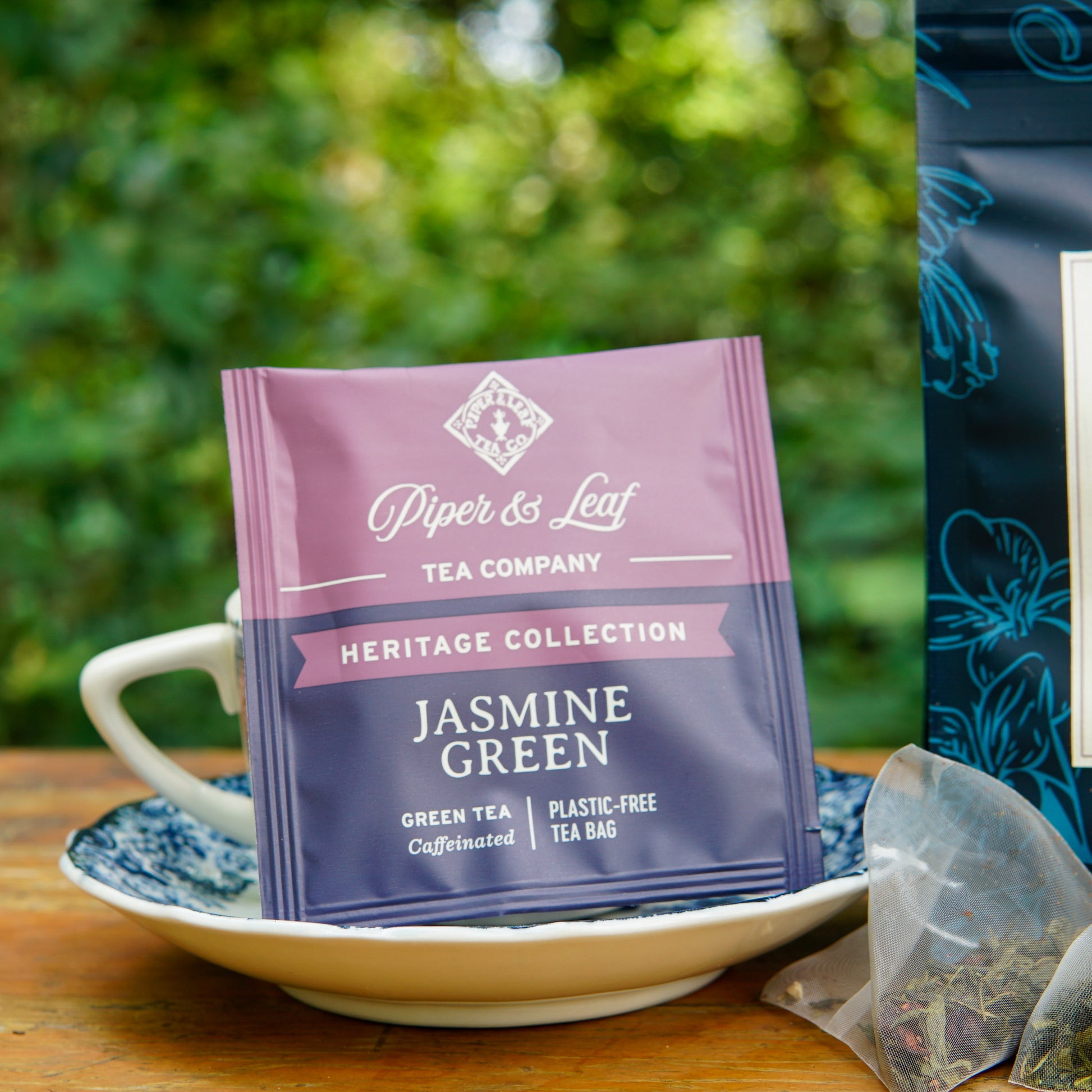 A Piper and Leaf Tea Co. Jasmine Green 50ct Individually Wrapped Tea Bag from the Heritage Collection sits upright in a teacup on a saucer, with greenery softly blurred in the background and a plastic-free tea bag open beside it.