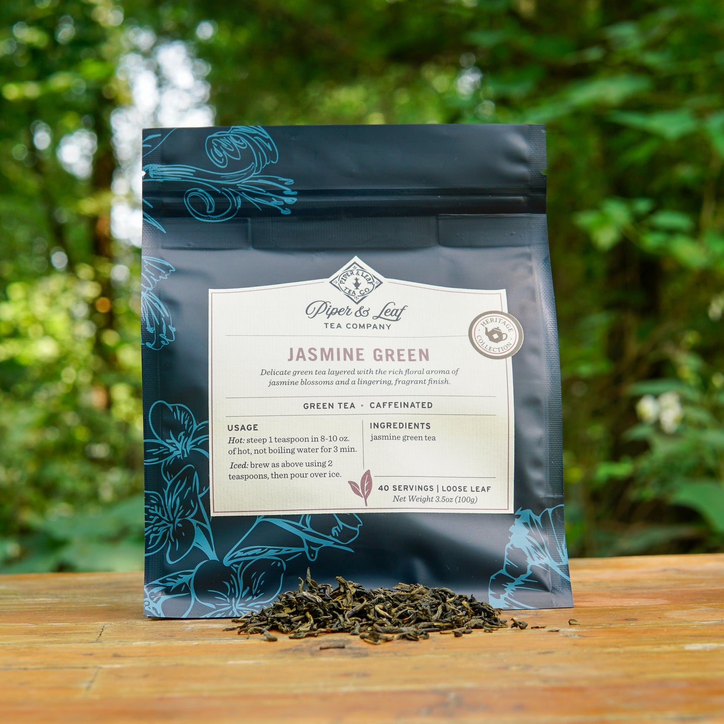 A pouch of Piper and Leaf Tea Co. Jasmine Green 40 Serving Loose Leaf tea from the Heritage Collection rests on a wooden surface outdoors, with loose tea leaves scattered in front of the package.