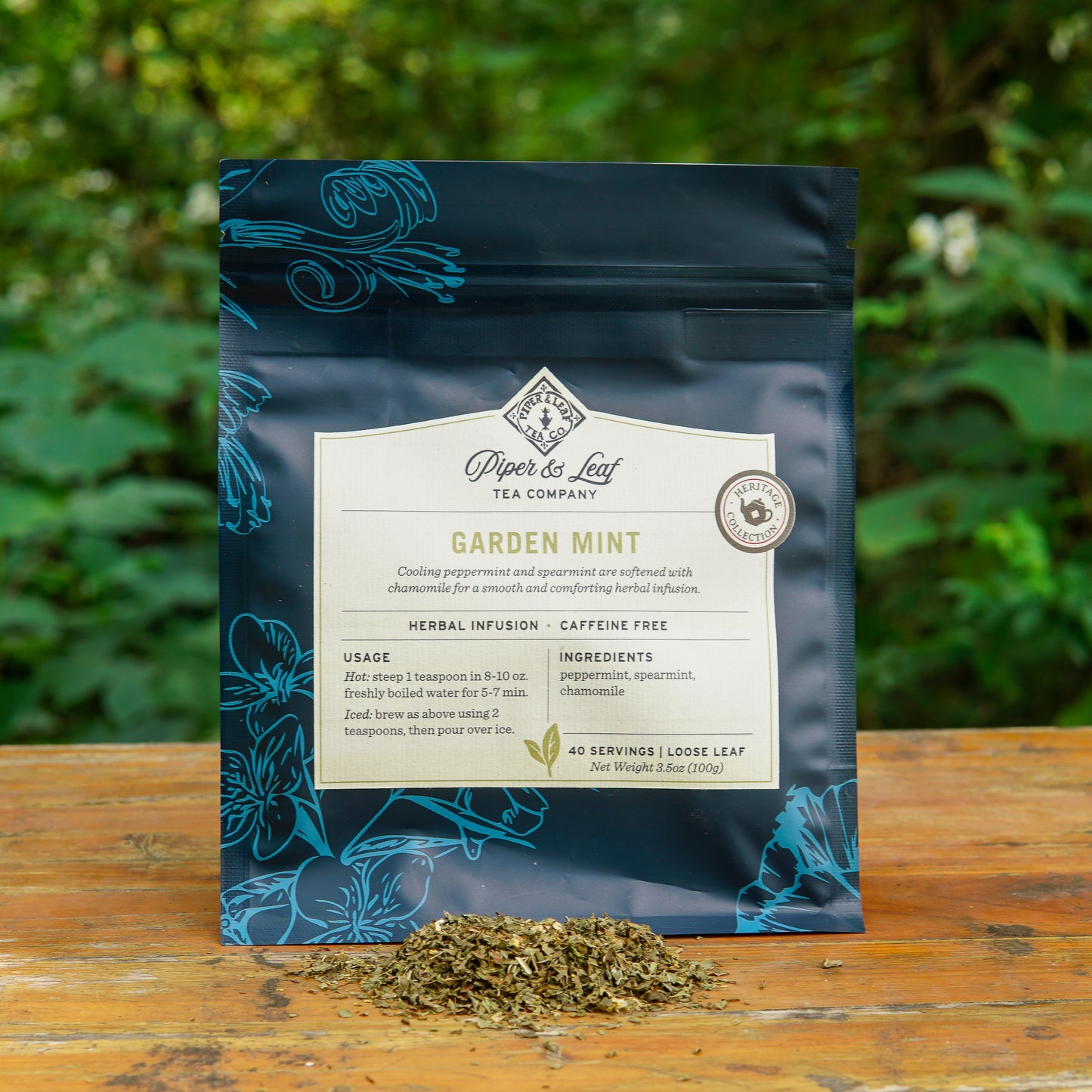 A blue bag of Piper and Leaf Tea Co. Garden Mint 40 Serving Loose Leaf tea rests on a wooden surface outdoors, with a small pile of this caffeine-free herbal infusion in front and lush green foliage in the background.