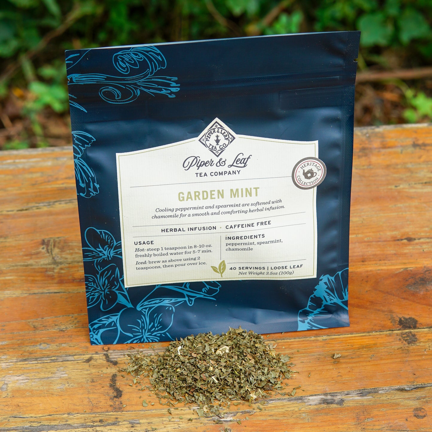 A bag of Piper and Leaf Tea Co. Garden Mint 40 Serving Loose Leaf rests on a wooden surface outdoors, with a small pile of the caffeine-free herbal infusion leaves displayed in front of the package.