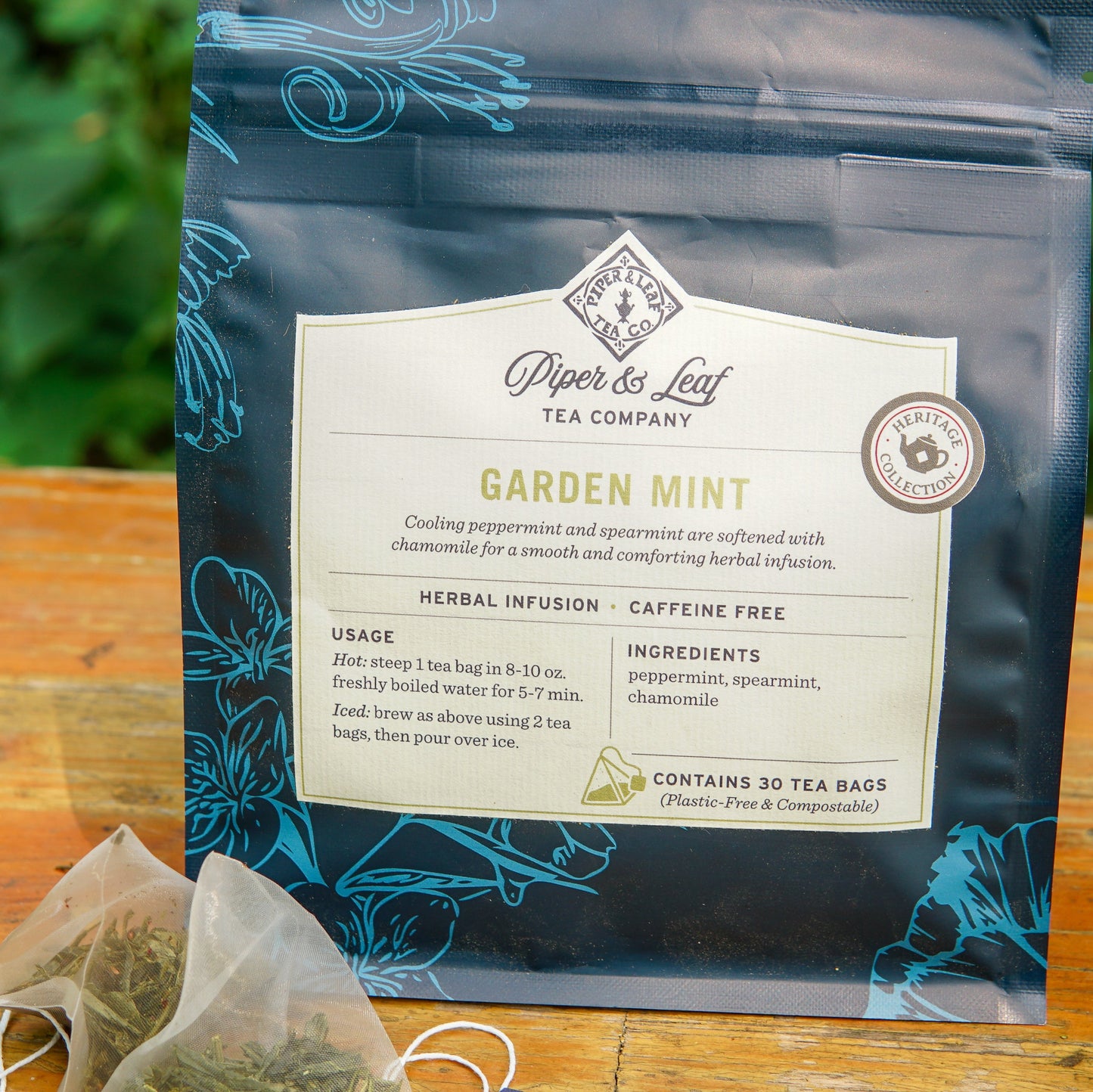 A bag of Piper and Leaf Tea Co. Garden Mint 30ct Tea Bags rests on a wooden surface, with two pyramid tea bags placed in front. The label details the ingredients and usage instructions, emphasizing that this caffeine-free blend provides a refreshing herbal infusion perfect for any time of day.