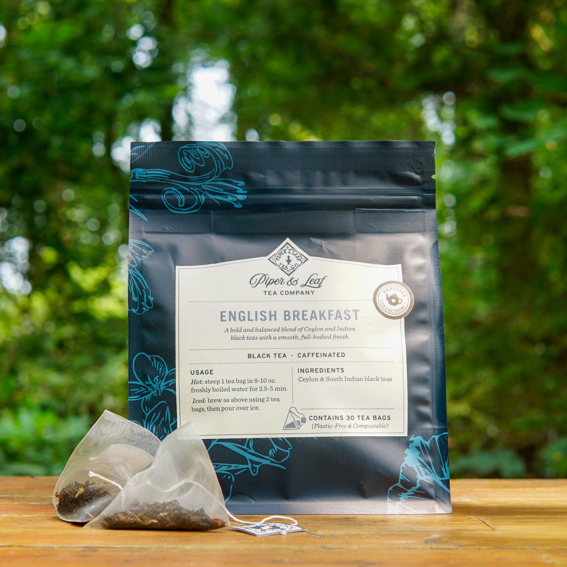 A package of Piper and Leaf Tea Co. English Breakfast 30ct Tea Bags from the Heritage Collection is displayed outdoors with two tea bags in front, set against a backdrop of greenery.