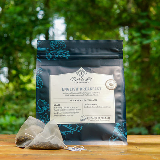A package of Piper and Leaf Tea Co. English Breakfast 30ct Tea Bags from the Heritage Collection is displayed outdoors with two tea bags in front, set against a backdrop of greenery.