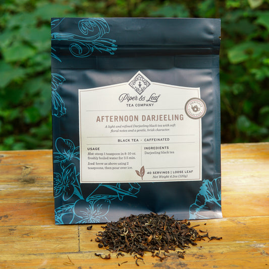 A bag of Piper and Leaf Tea Co. Afternoon Darjeeling 40 Serving Loose Leaf from the Heritage Collection rests outdoors on a wooden surface, with some loose-leaf tea thoughtfully scattered in front of it.