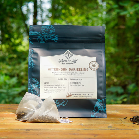A pouch of Piper and Leaf Tea Co. Afternoon Darjeeling 30ct Tea Bags rests on a wooden surface outdoors, with two plastic-free tea bags and loose tea leaves displayed in front of the package.
