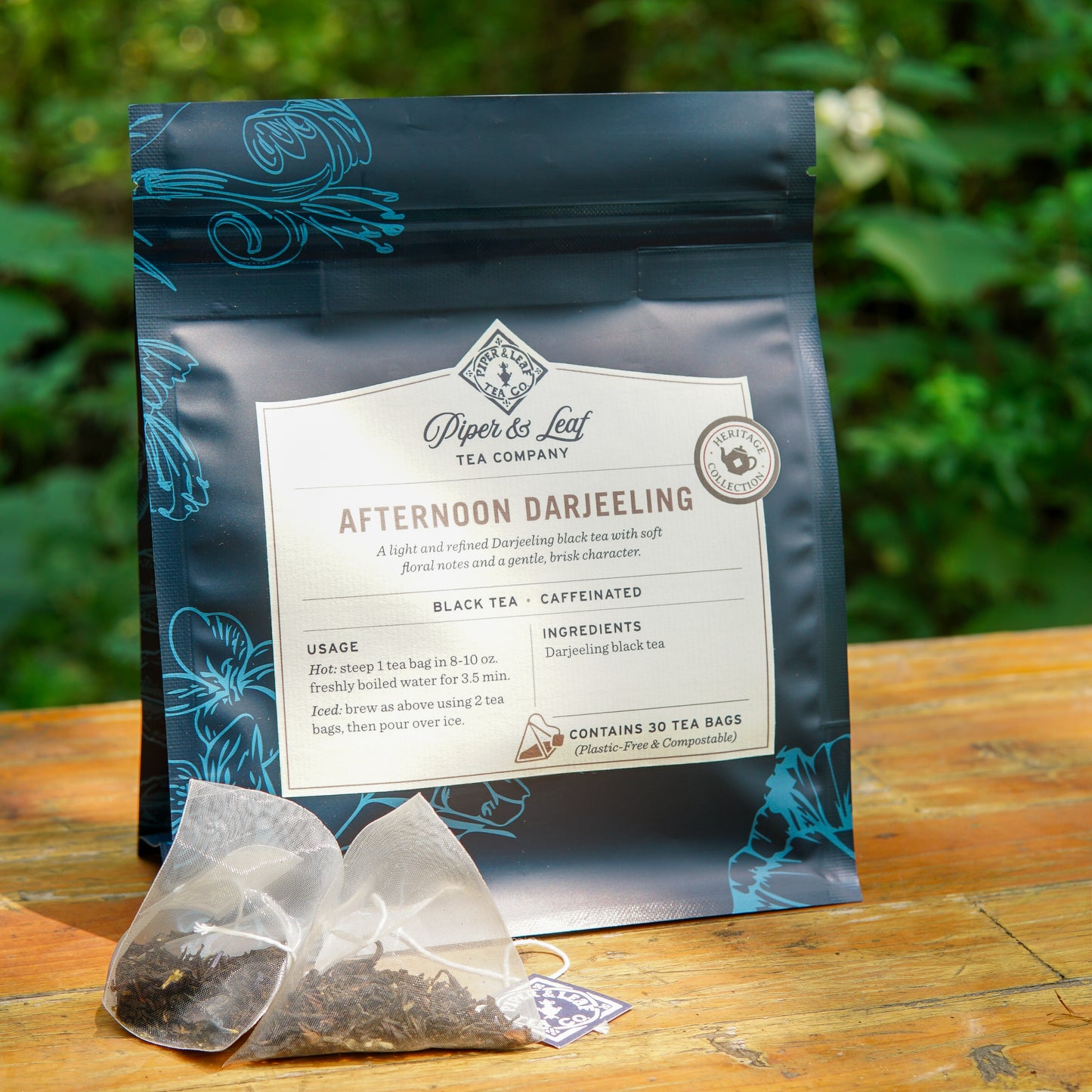 A blue package of Piper and Leaf Tea Co. Afternoon Darjeeling 30ct Tea Bags from the Heritage Collection rests on a wooden surface outdoors, with two plastic-free tea bags and loose Darjeeling black tea displayed in front.