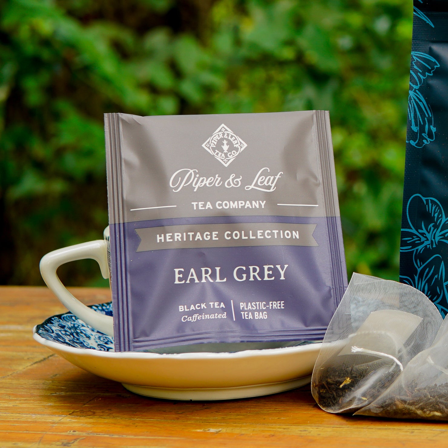 A box of Piper and Leaf Tea Co. Earl Grey 50ct Individually Wrapped Tea Bags rests on a saucer next to a cup and a used tea bag, with greenery softly blurred in the background.