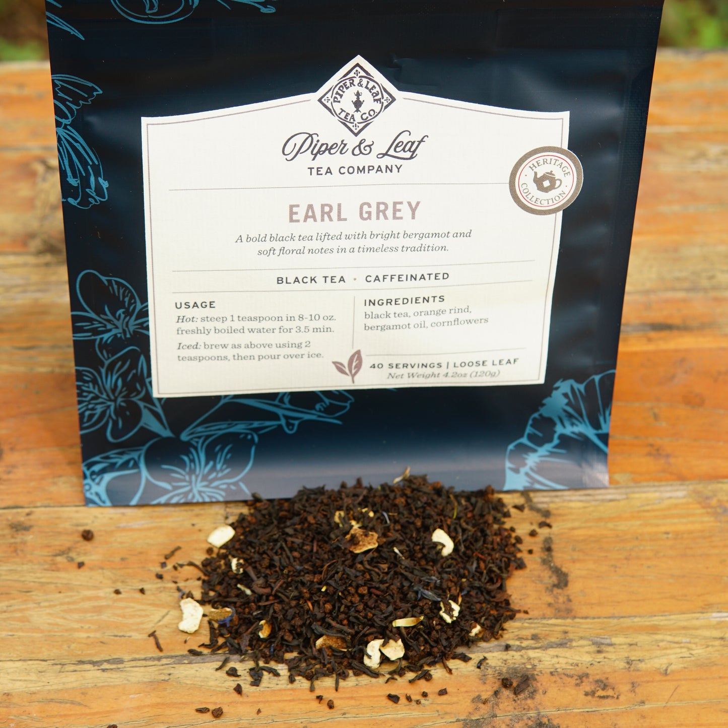 A bag of Piper and Leaf Tea Co. Earl Grey 40 Serving Loose Leaf Tea stands behind a small pile of loose leaf tea on a wooden surface outdoors.