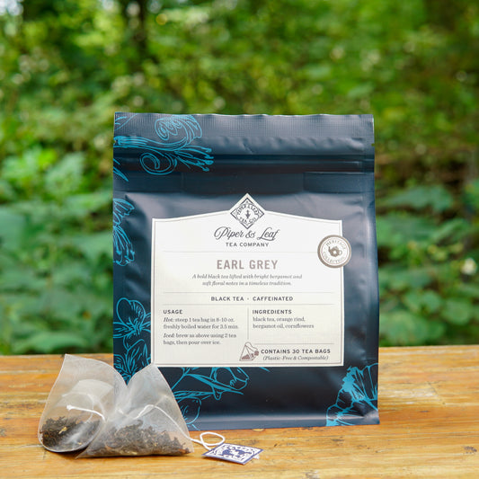 A box of Piper and Leaf Tea Co. Earl Grey 30ct Tea Bags from the Heritage Collection is displayed on a wooden surface, with two black tea bags in front and greenery in the background.