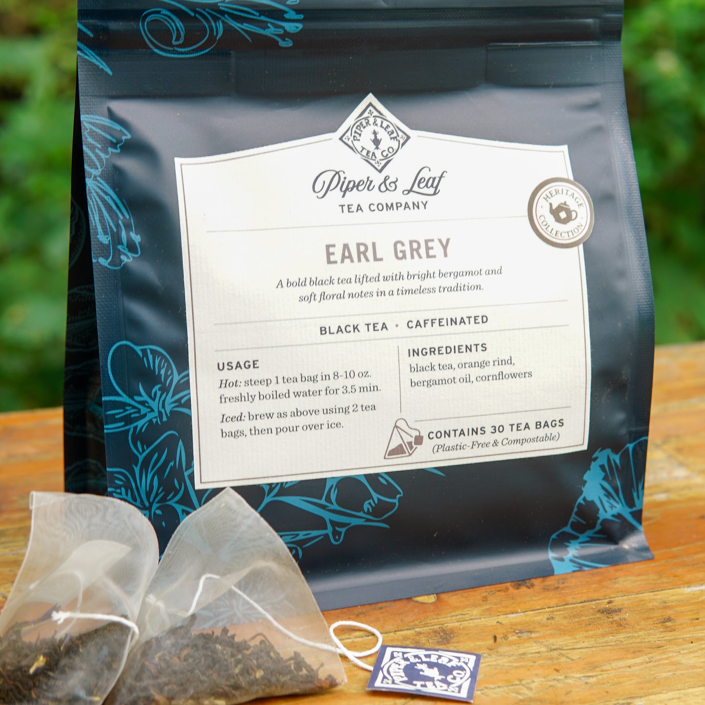 A box of Piper and Leaf Tea Co. Earl Grey 30ct Tea Bags from the Heritage Collection is displayed with two tea bags and loose tea leaves in front on a wooden surface.