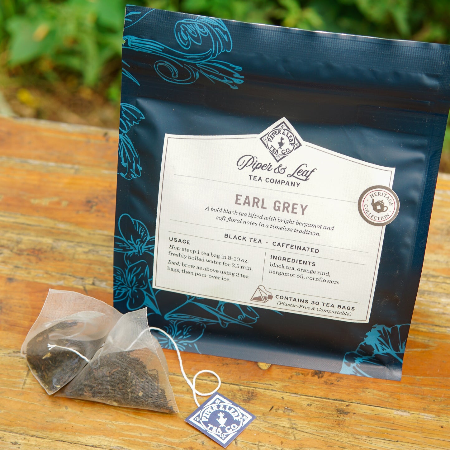 A box of Piper and Leaf Tea Co. Earl Grey 30ct Tea Bags from the Heritage Collection rests on a wooden surface beside a single pyramid tea bag filled with loose black tea.