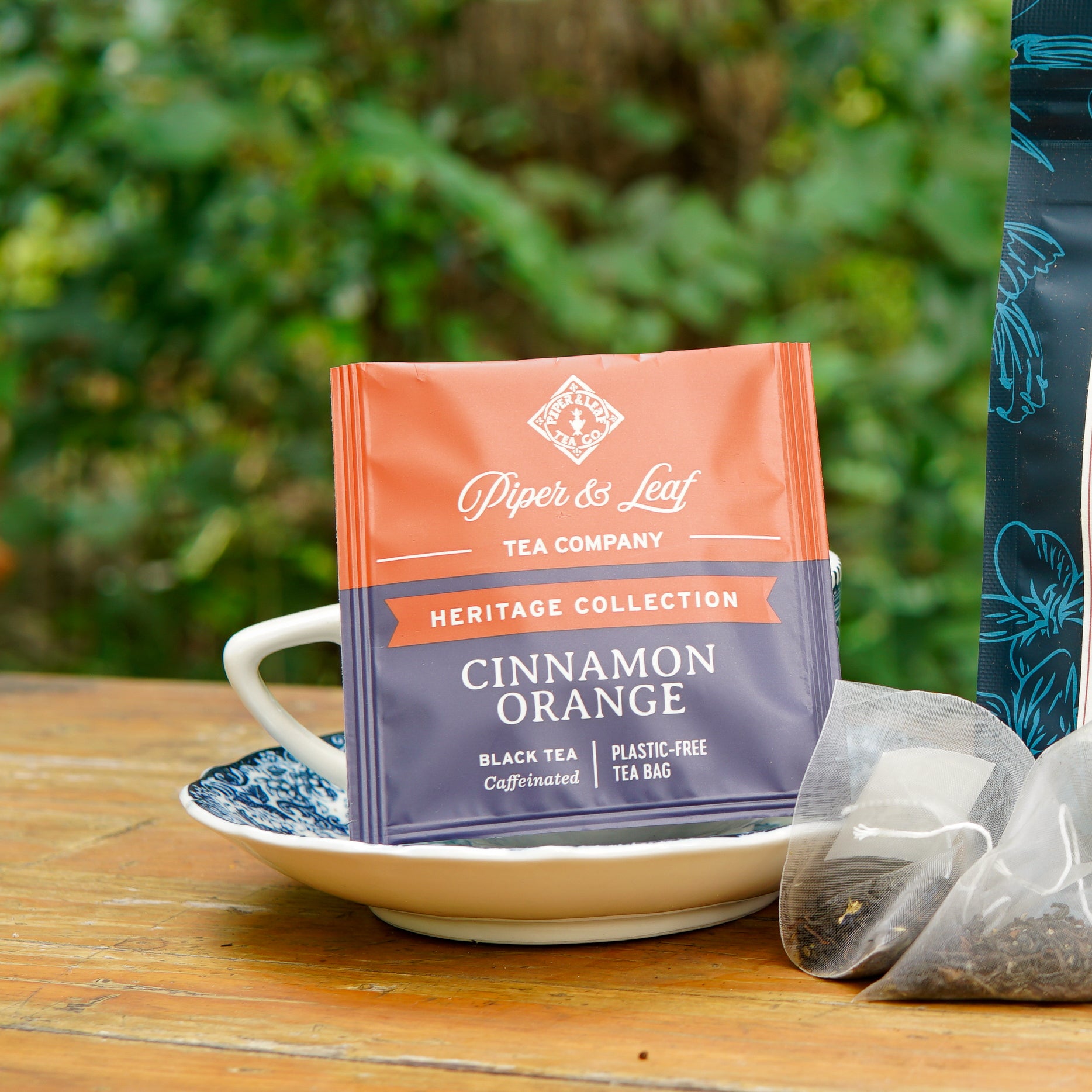 A Piper and Leaf Tea Co. Cinnamon Orange 50ct Individually Wrapped Tea Bag from the Heritage Collection rests on a white and blue saucer, accompanied by a tea cup and another black tea bag, all arranged on a wooden outdoor table.