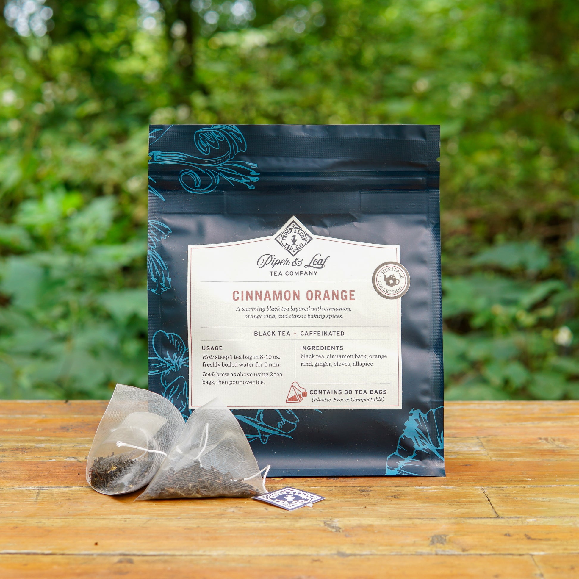 A box of Piper and Leaf Tea Co. Cinnamon Orange 30ct Tea Bags is displayed on a wooden surface outdoors, with two pyramid tea bags placed in front of it.