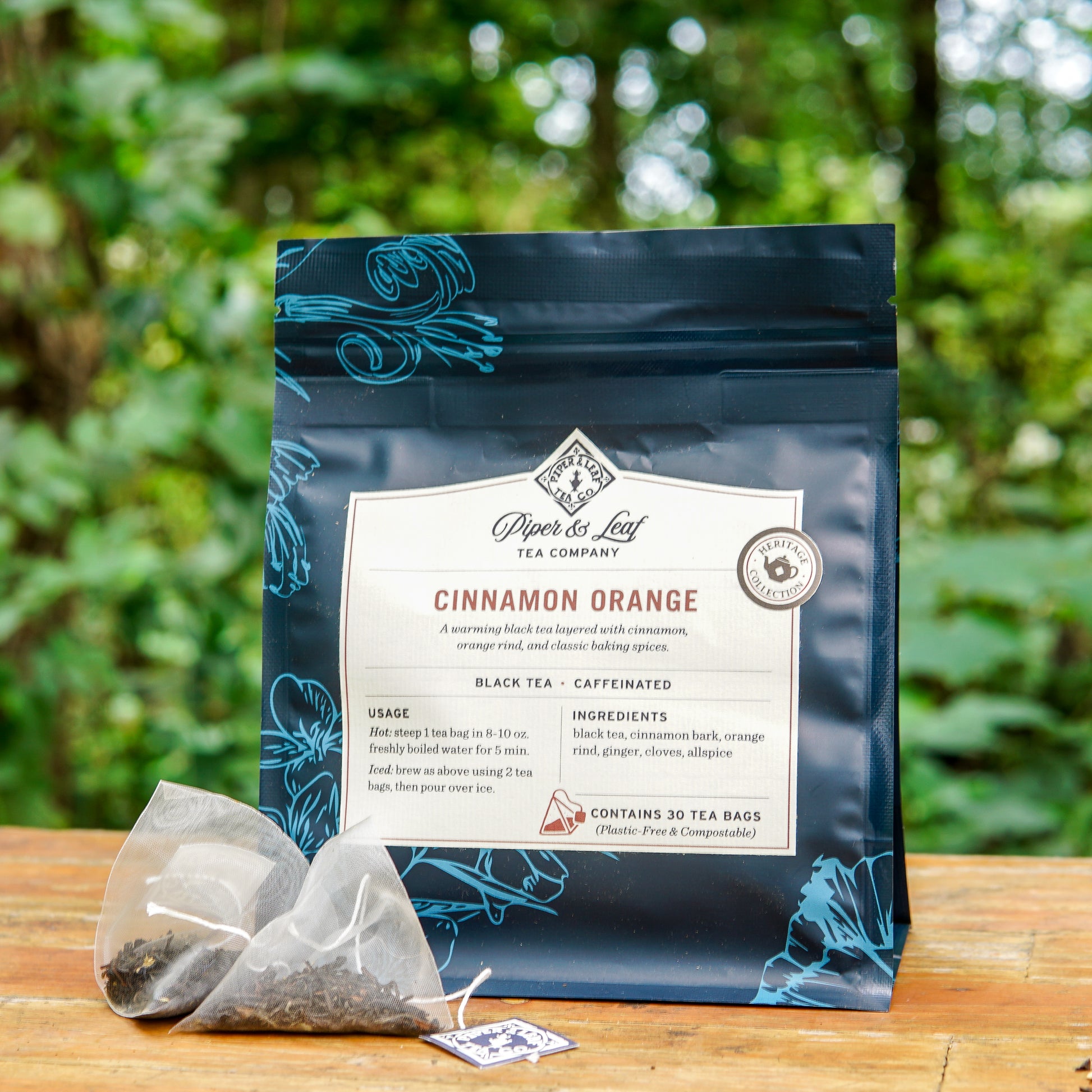 A bag of Piper and Leaf Tea Co. Cinnamon Orange 30ct Tea Bags rests on a wooden surface outdoors, with two pyramid tea bags placed in front of the package and a forest background visible.