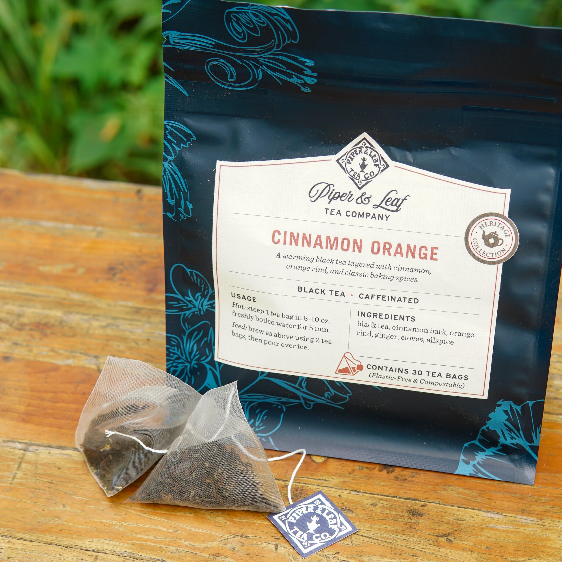 A package of Piper and Leaf Tea Co. Cinnamon Orange 30ct Tea Bags rests on a wooden surface, with two pyramid-shaped tea bags displayed in front of it.