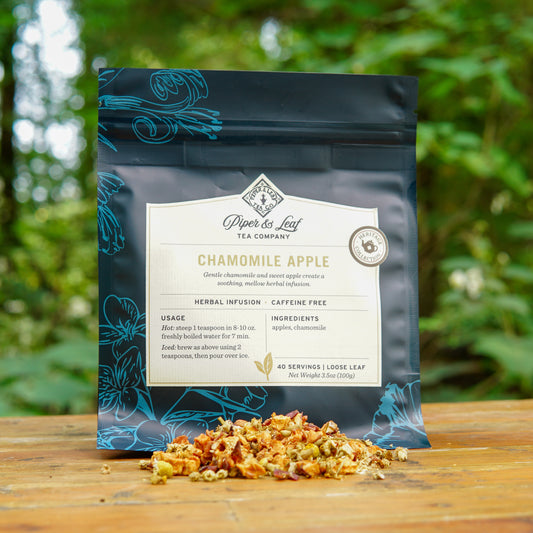 A bag of Piper and Leaf Tea Co.’s Chamomile Apple 40 Serving Loose Leaf Tea rests on a wooden surface outdoors, with dried tea leaves scattered in front, highlighting this soothing caffeine-free herbal infusion.